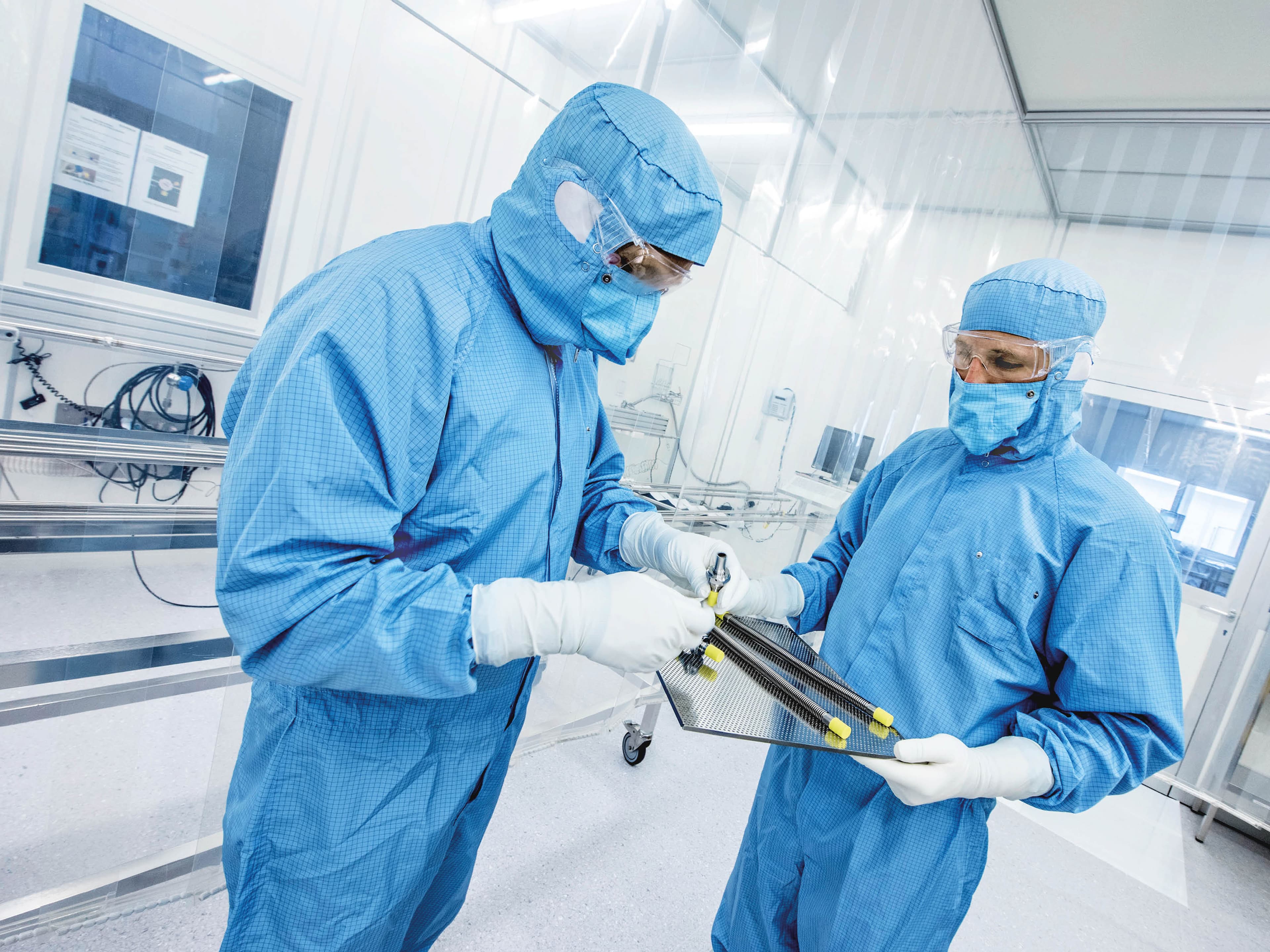 Two people in cleanroom clothing holding various corrugated stainless steel hoses on a tray