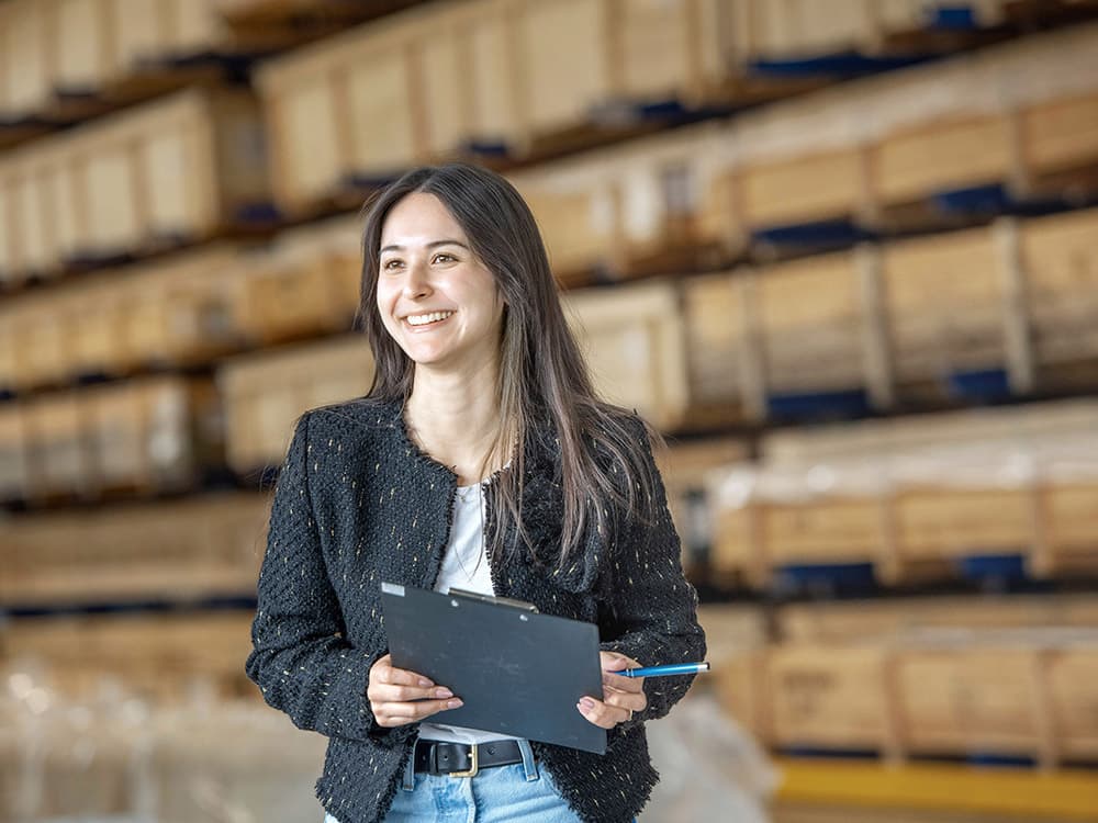 Woman with clipboard and pen in hand, smiling in front of a high-bay warehouse with crates