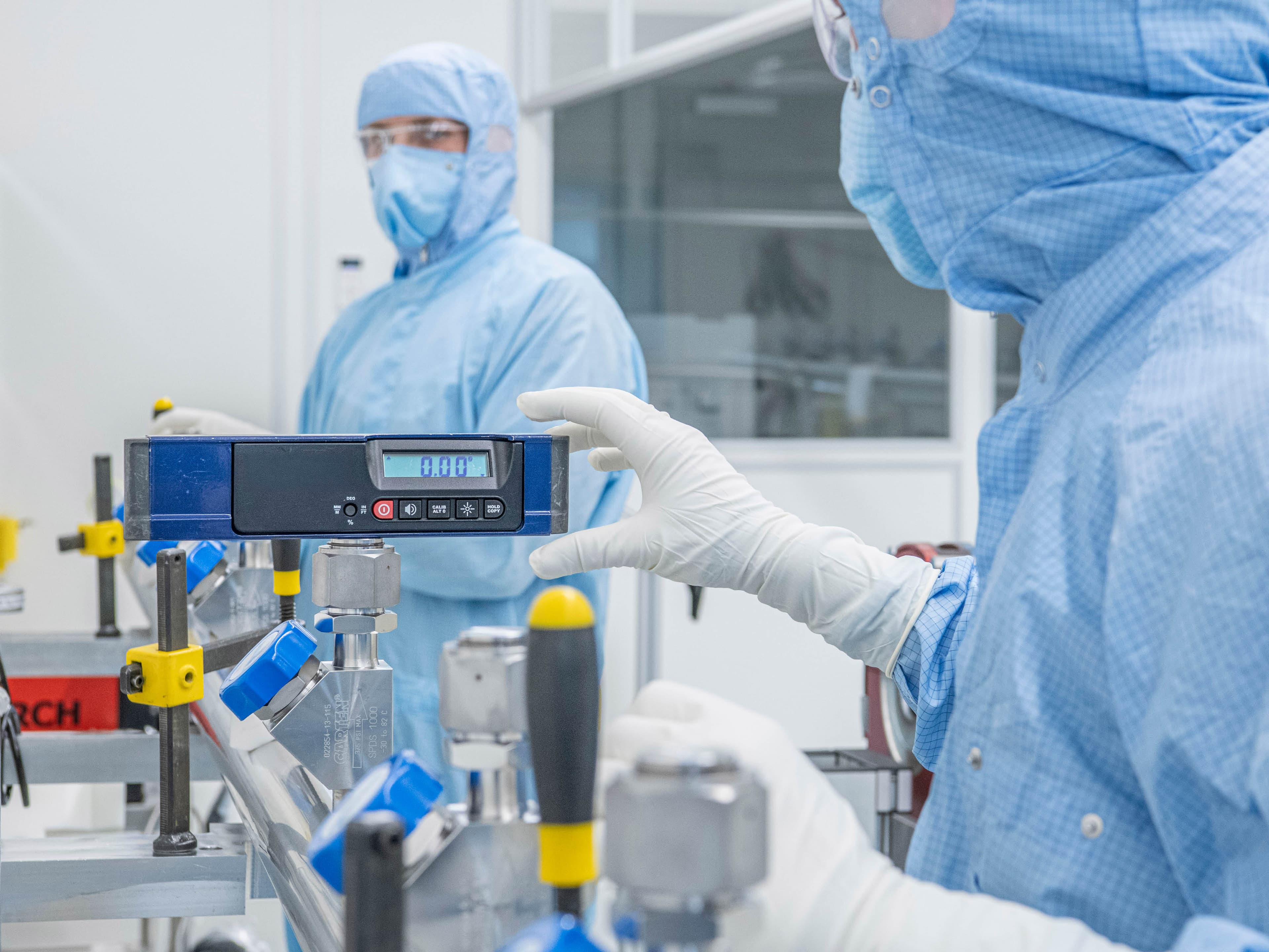 Two employees in cleanroom protective clothing are working on a manifold and checking values.