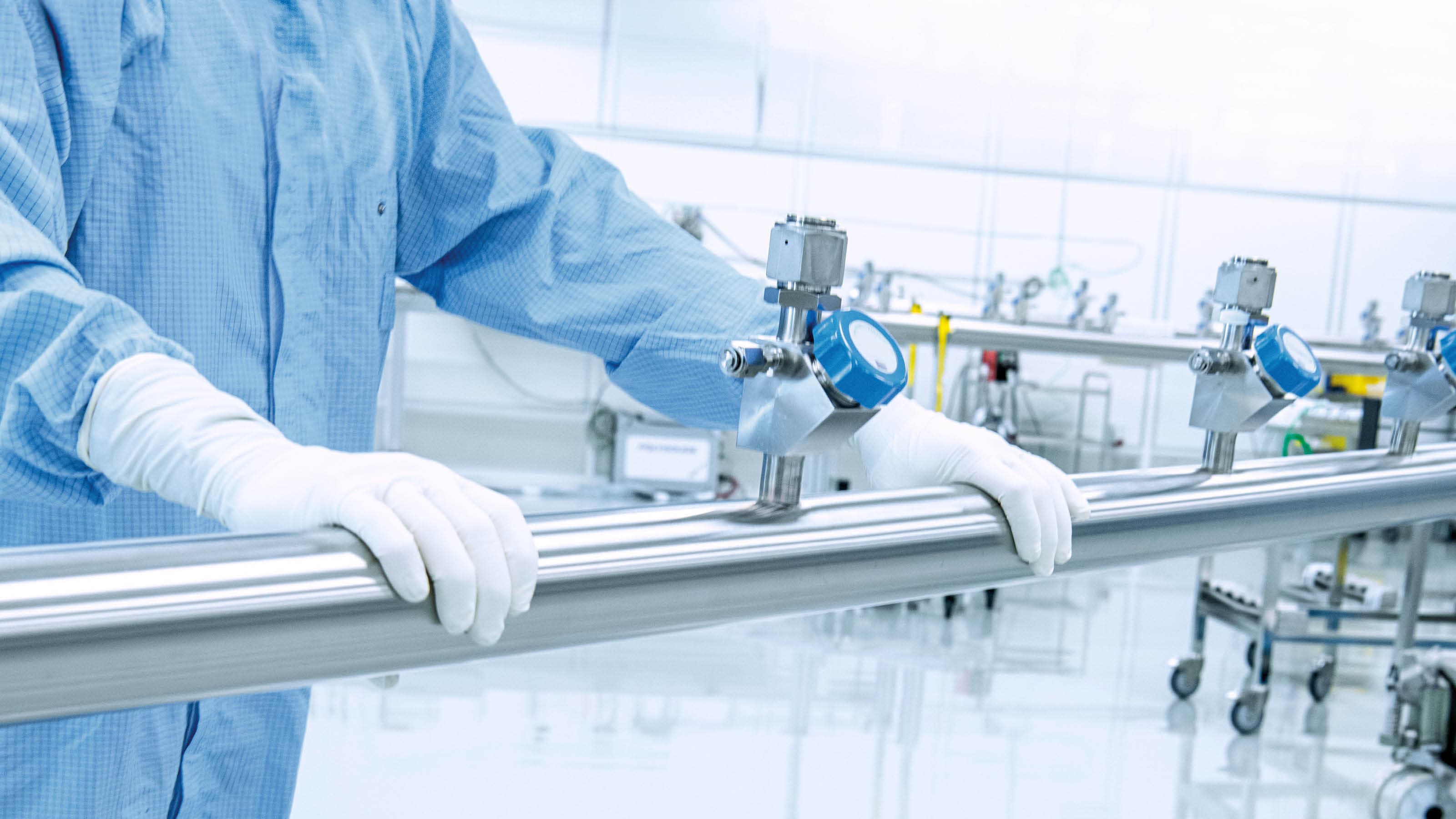 An employee wearing cleanroom protective clothing works at a manifold. The cleanroom and the equipment can be clearly seen in the background.