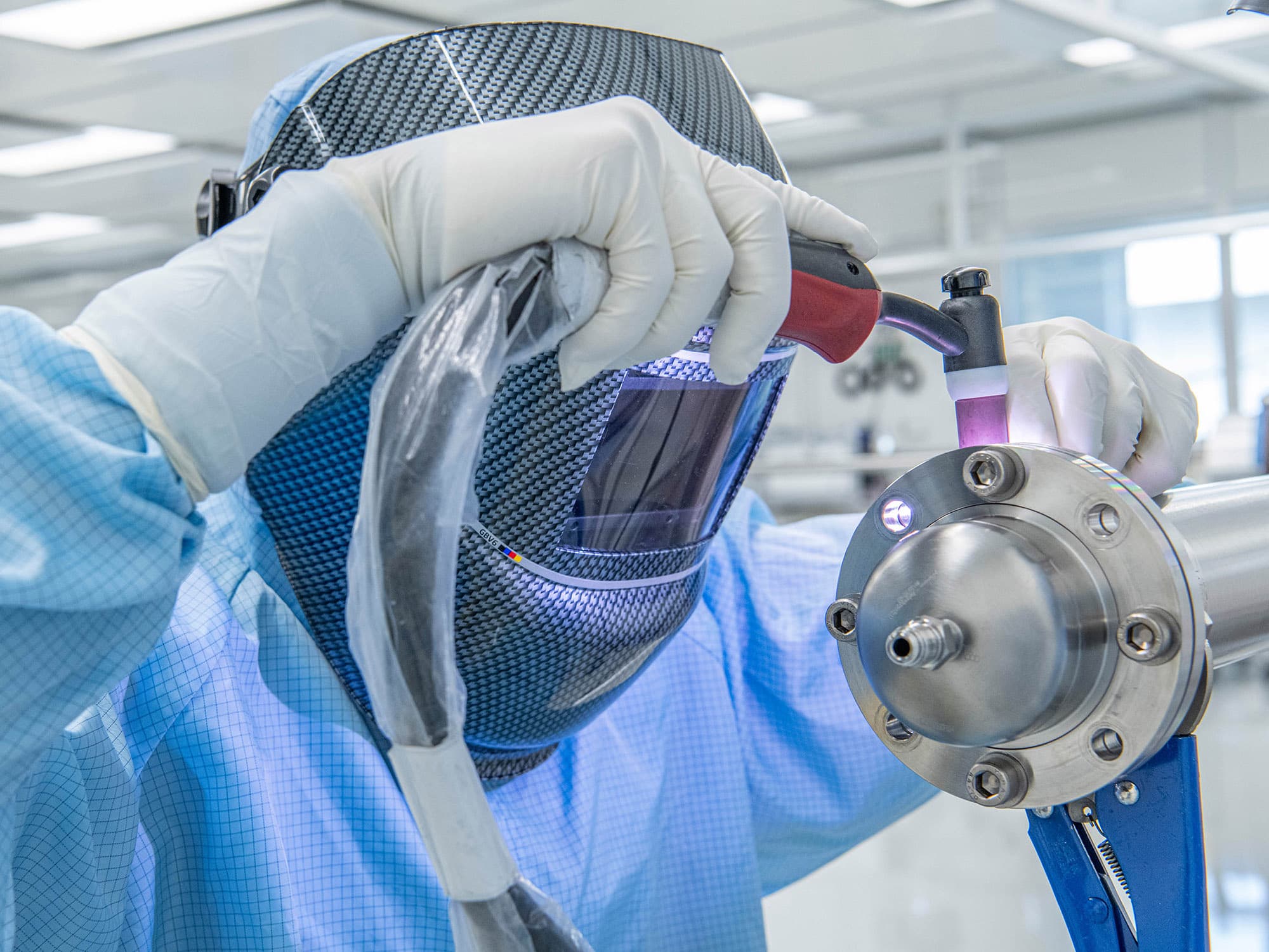 Welders in protective clothing and helmets manually weld a manifold in a clean room.