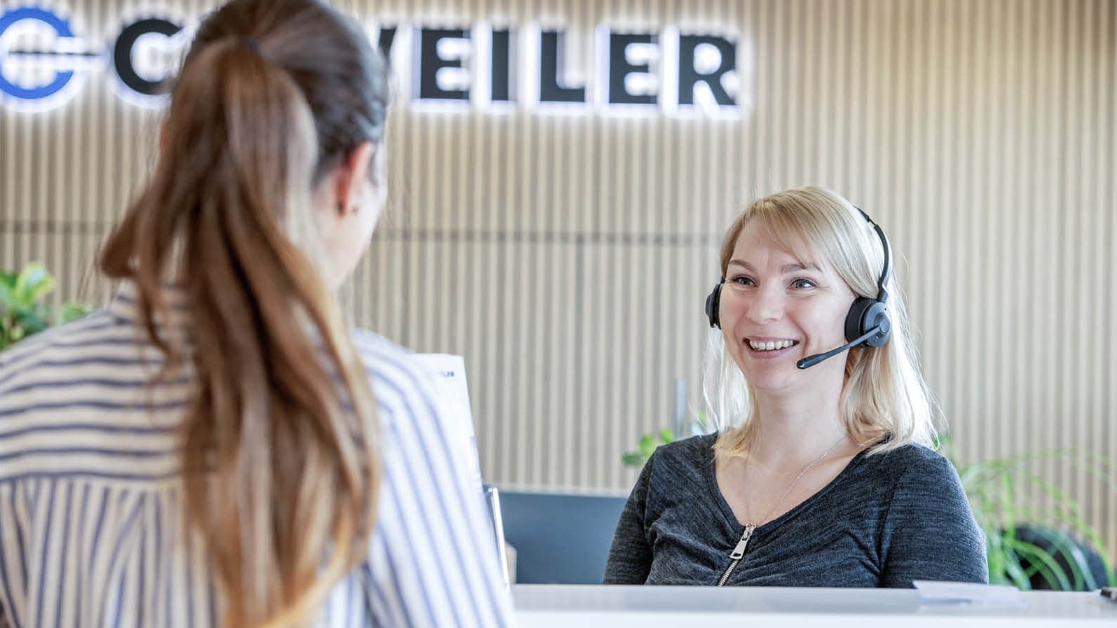 A receptionist with a headset speaks to a visitor; wooden paneling and the Dockweiler logo can be seen in the background.
