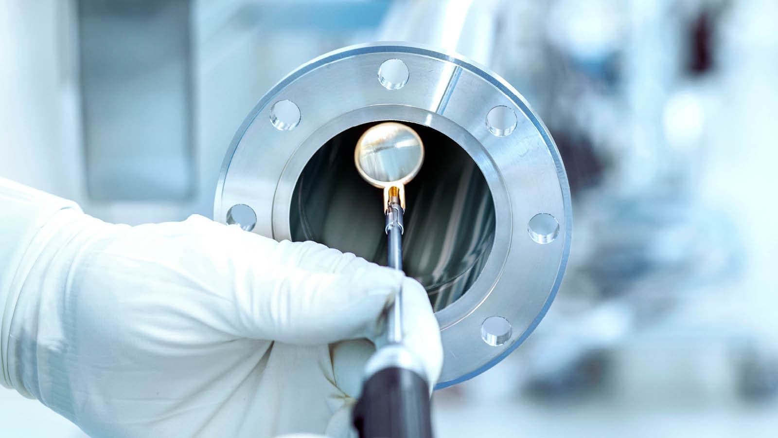 A hand in white latex gloves uses a hand mirror to check the weld of a component